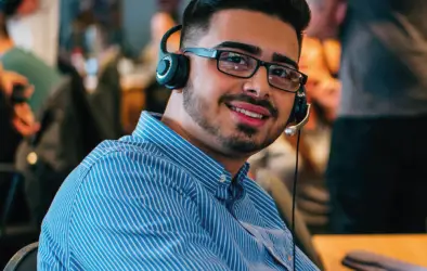 A young man wearing glasses and a blue striped shirt smiles at the camera while wearing a headset in a busy office environment, surrounded by lively conversations and teaser discussions for an upcoming project carousel.
