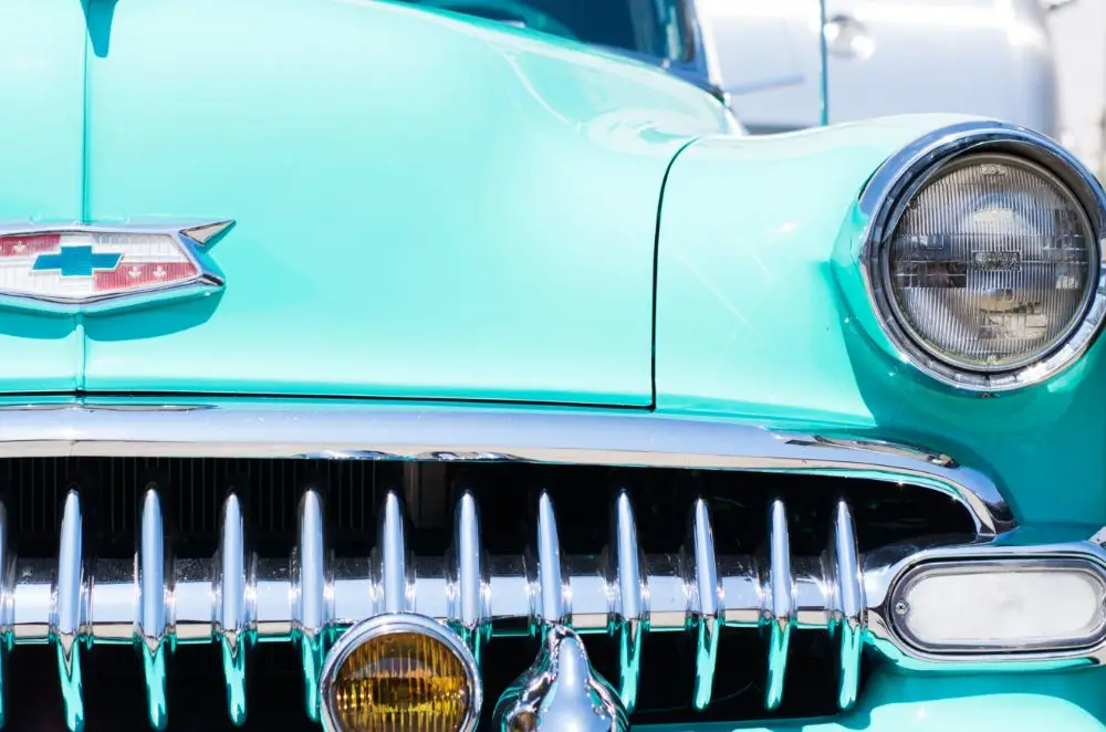 Close-up of the front of a vintage turquoise Chevrolet, showing its chrome grille, headlamp, and Chevrolet emblem—a perfect example for those seeking American classic car insurance.
