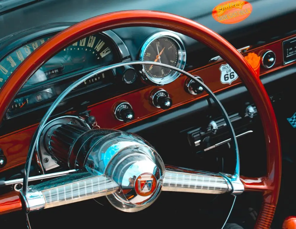 Close-up of a classic car’s dashboard and steering wheel, featuring retro gauges, chrome details, a Route 66 badge, and a red-orange Summer Roadster sticker—perfect for an American classic car insurance advert.