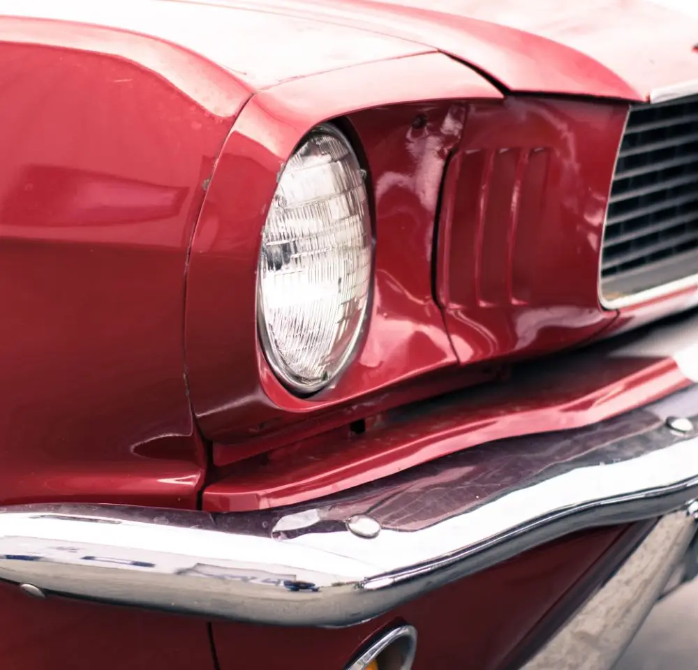 Close-up of the front left side of a vintage red car, featuring its round headlamp, chrome bumper, and classic bonnet lines—a perfect example for those seeking American classic car insurance.