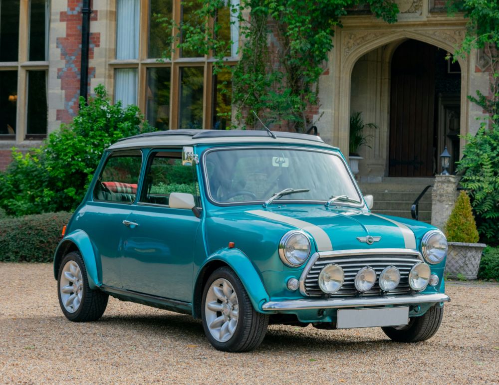 Classic teal Mini Cooper with white racing stripes parked on a gravel driveway in front of an elegant historic building with arched doorways and tall windows, surrounded by lush greenery.