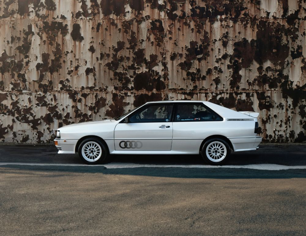 White classic Audi Quattro coupe with rear spoiler and rally decals parked in front of a rusted industrial metal wall, highlighting the car’s vintage design against a gritty urban backdrop.