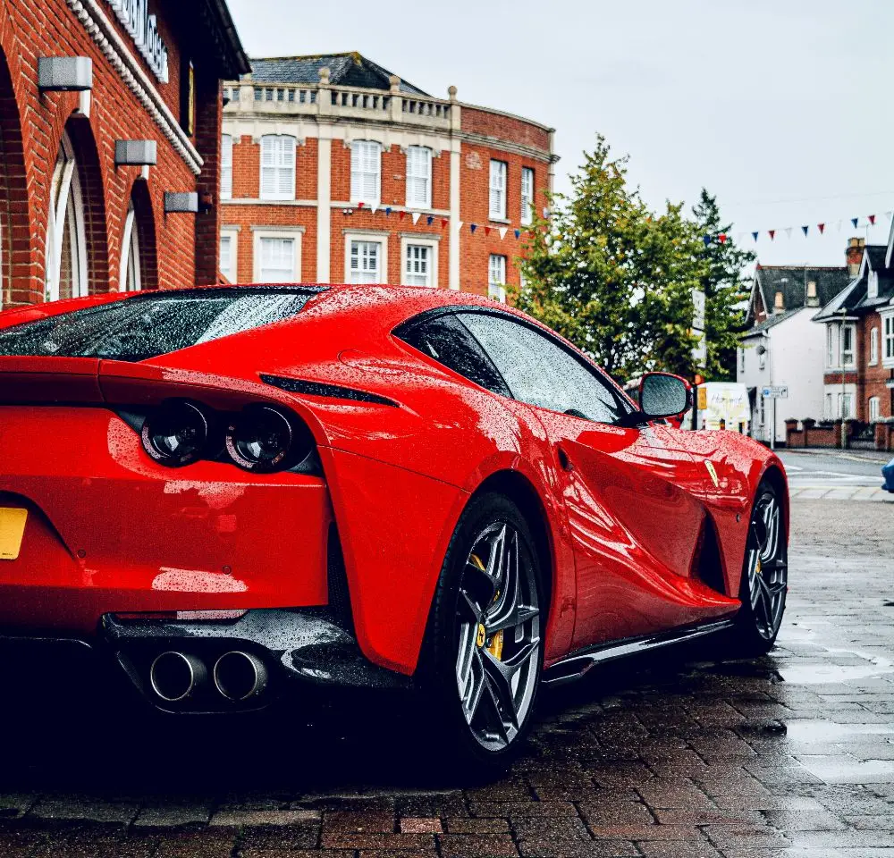 A shiny red Ferrari parked on a wet brick street, reflecting raindrops. Red brick buildings and trees are visible in the background on a cloudy day—a perfect scene to consider Ferrari insurance for your prized car.