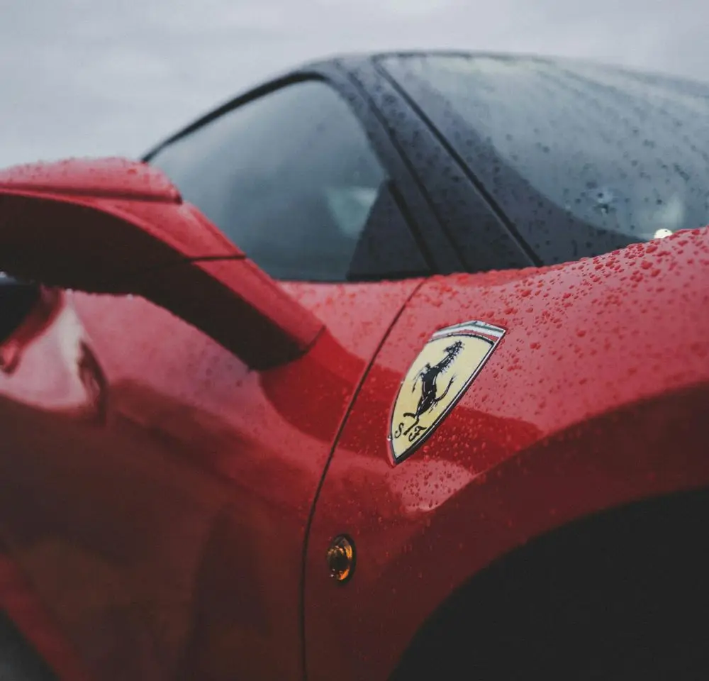Close-up of a red Ferrari sports car with rain droplets on the surface. The Ferrari logo is visible on the side, highlighting why exclusive Ferrari insurance is essential. A cloudy sky is reflected on the car’s window.