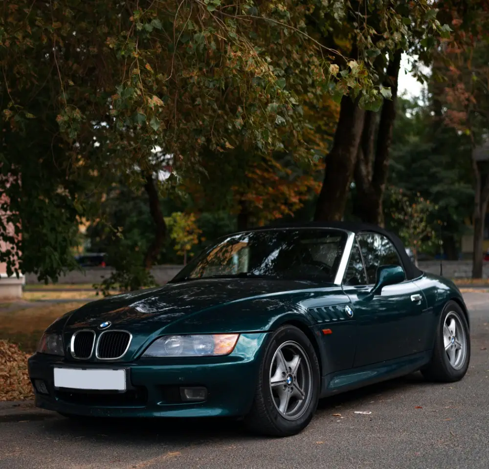 A dark green BMW Z3 convertible is parked on a quiet street under leafy trees, fallen leaves scattered around. The scene highlights the beauty of autumn and the allure of owning a BMW Z3—perfect for those considering BMW Z3 insurance.
