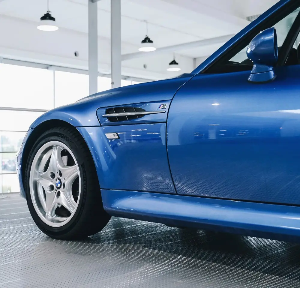 Close-up of the front side of a blue BMW Z3 sports car parked indoors, showing the front wheel, wing mirror, and M badge on the wing, with large windows and ceiling lights in the background—perfect for classic car insurance.