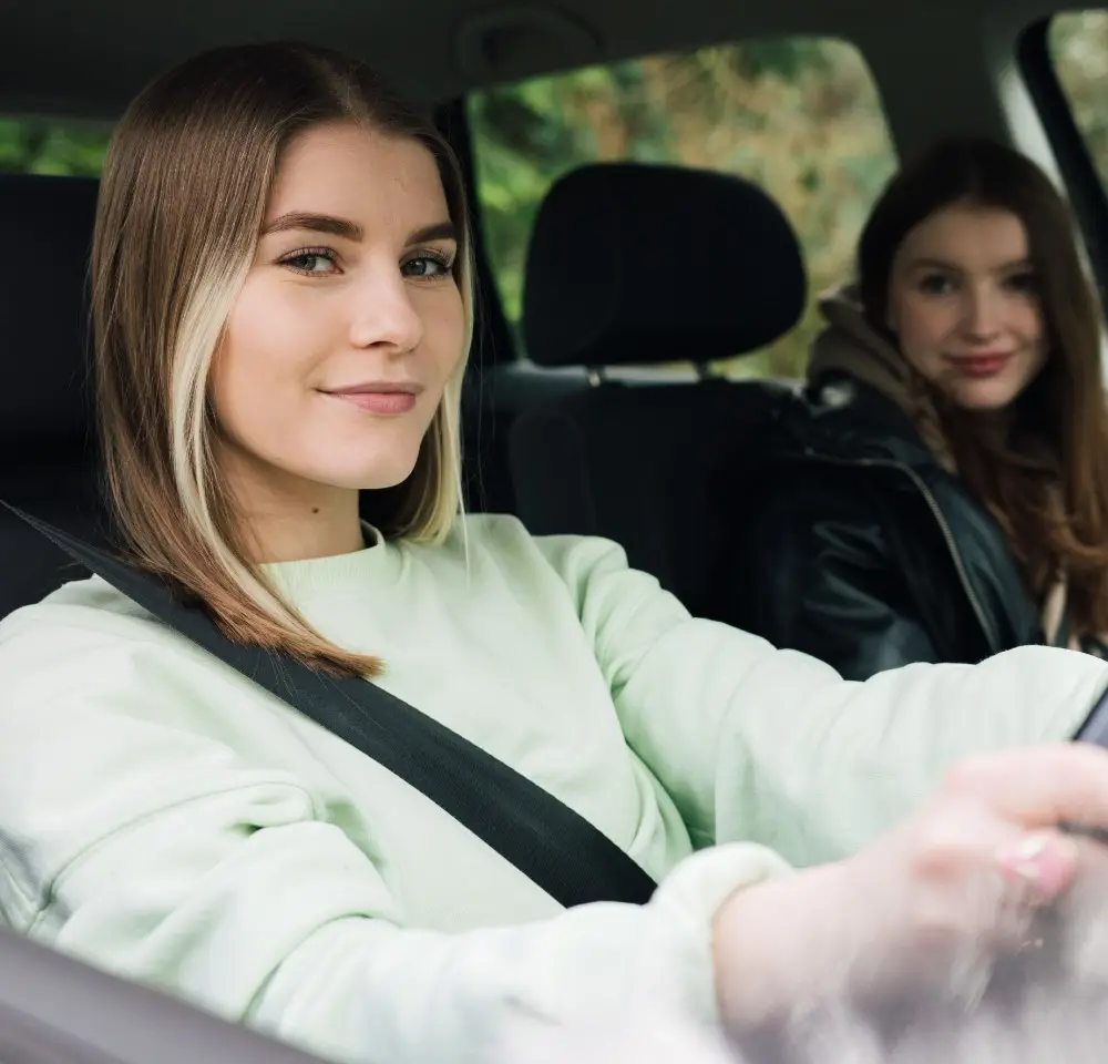 Two young women sit in a car; the driver smiles at the camera from behind the wheel, whilst her friend in the back seat grins too—both enjoying their ride, possibly protected with BMW Z3 insurance for peace of mind on every journey.