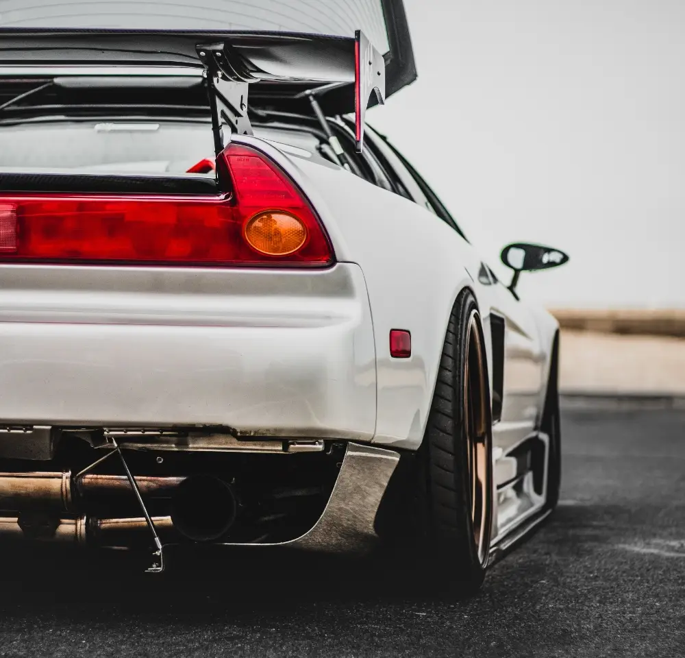 A low-angle view of the rear of a white sports car with a large rear wing, custom exhaust, and wide tyres, parked on tarmac. This sleek design with red tail lights and aerodynamic parts might turn heads—and could impact your Honda insurance.