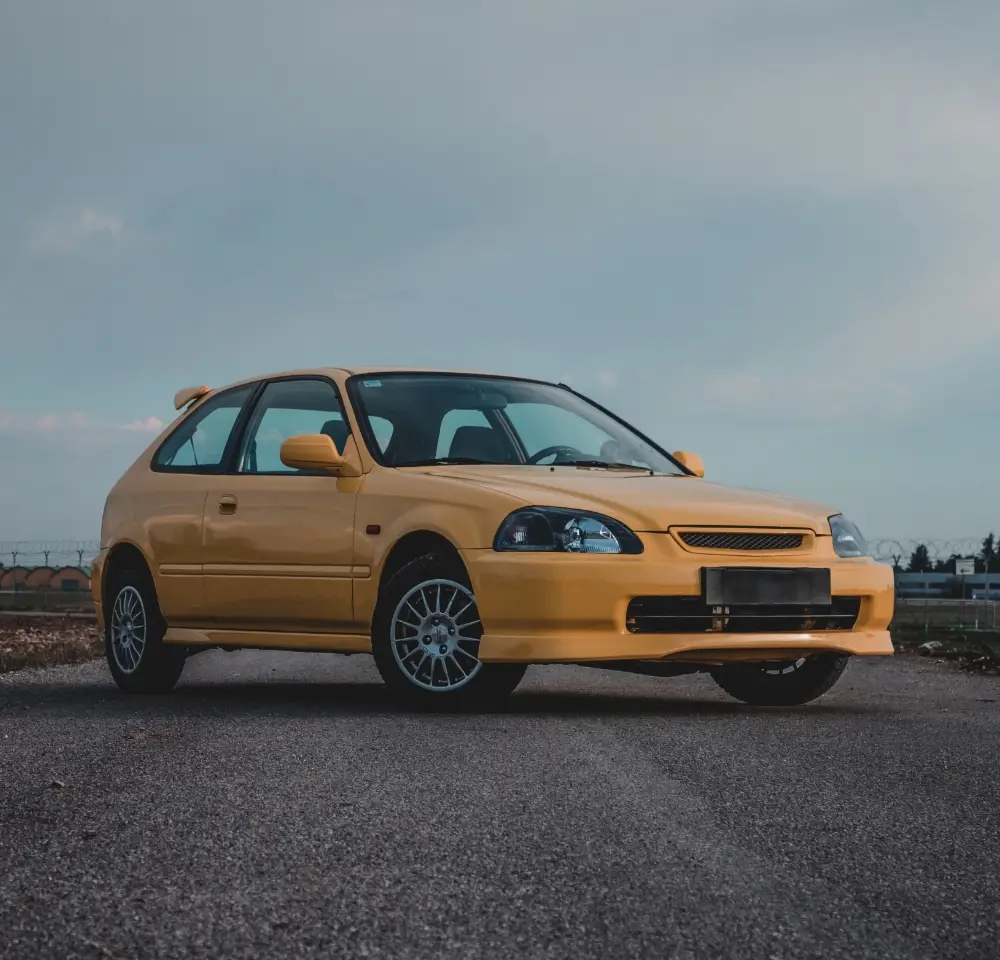 A yellow two-door car is parked on a paved surface outdoors, with a cloudy sky in the background. The clean, well-maintained vehicle sits at an angle showing its front and side—ideal for showcasing Honda insurance options.