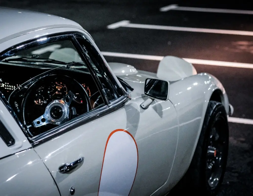 Close-up of a vintage white Lotus Elan parked in an empty car park at night, focusing on the driver’s side window, steering wheel, and bonnet with a racing number outlined in orange—perfect for classic car insurance enthusiasts.