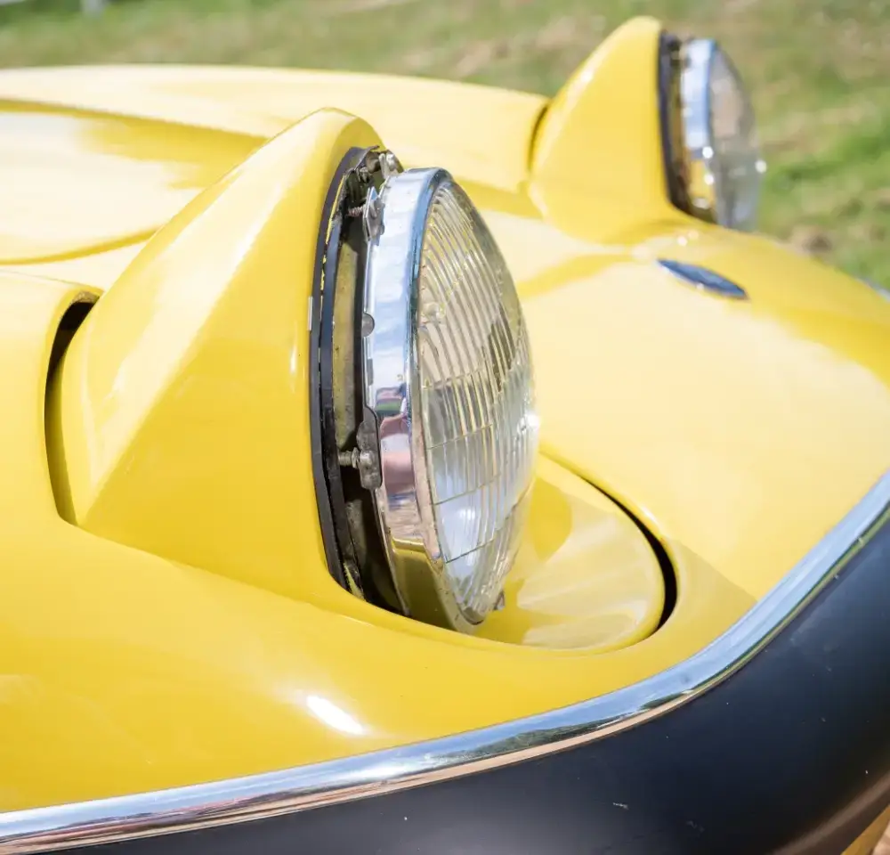 Close-up of a yellow Lotus Elan’s distinctive pop-up headlamps, lights raised. The car’s curved bonnet and part of the windscreen are visible, set against a grassy background—perfect for a classic car insurance advert.