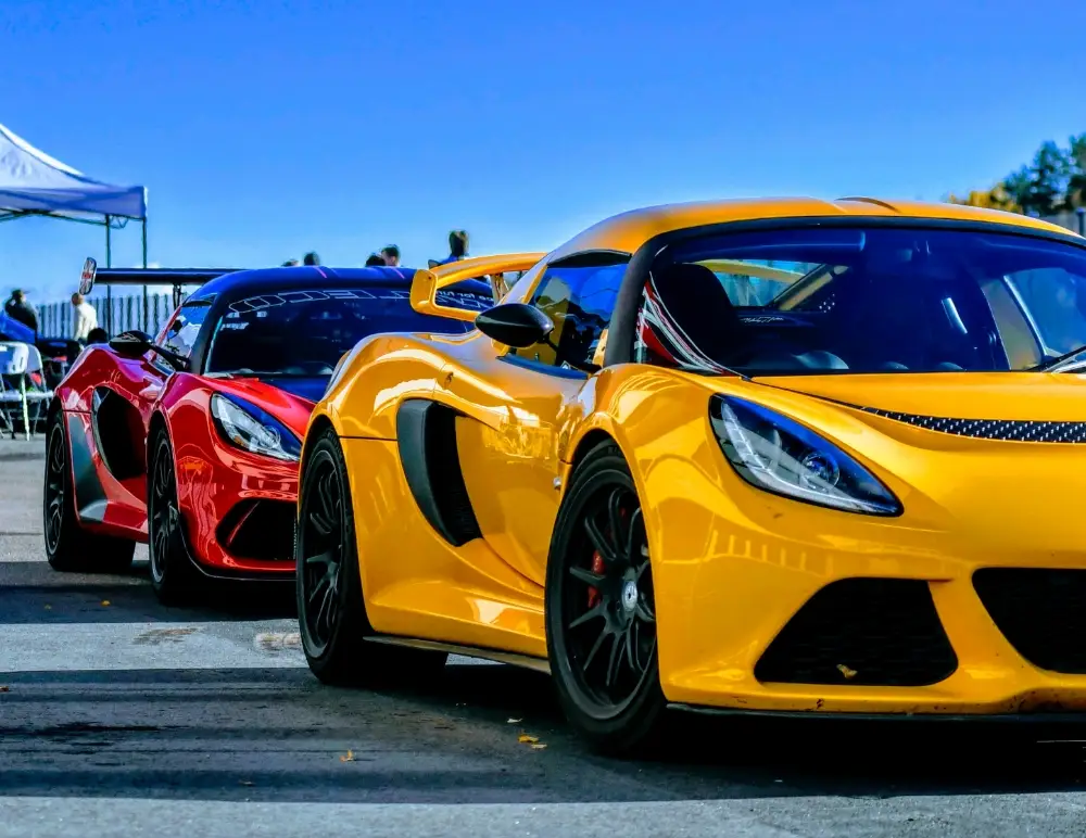 Two sleek sports cars, one yellow and one red, are parked side by side under a clear blue sky at an outdoor event, with people and a marquee visible in the background—perfect for discussing Lotus Elise car insurance.