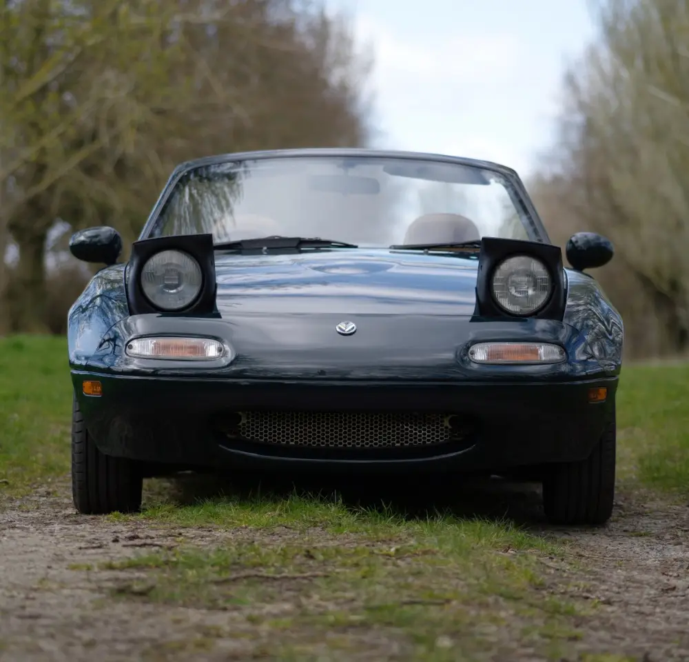 A front view of a dark green Mazda MX-5 convertible with its pop-up headlights raised, parked on a grassy, tree-lined path under a cloudy sky—a timeless Mazda showcasing iconic style and adventure.