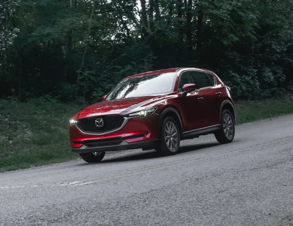A red Mazda SUV drives on a tarmacked road bordered by dense green trees and foliage, its headlights on during daylight—a reminder to keep your car insurance up to date for every journey.