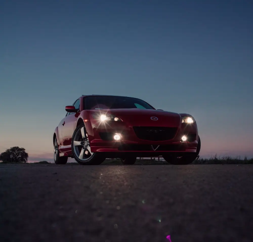 A red Mazda sports car with headlights on is parked on an empty road at dusk, with a clear sky and faint trees visible in the background.