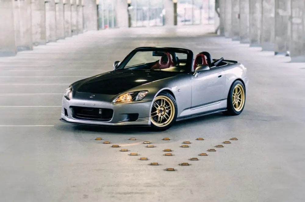 A silver and black Honda S2000 convertible with gold wheels, known for unique Honda S2000 insurance rates, is parked in an empty, covered car park. The car faces forward amid concrete columns and natural light streaming from behind.