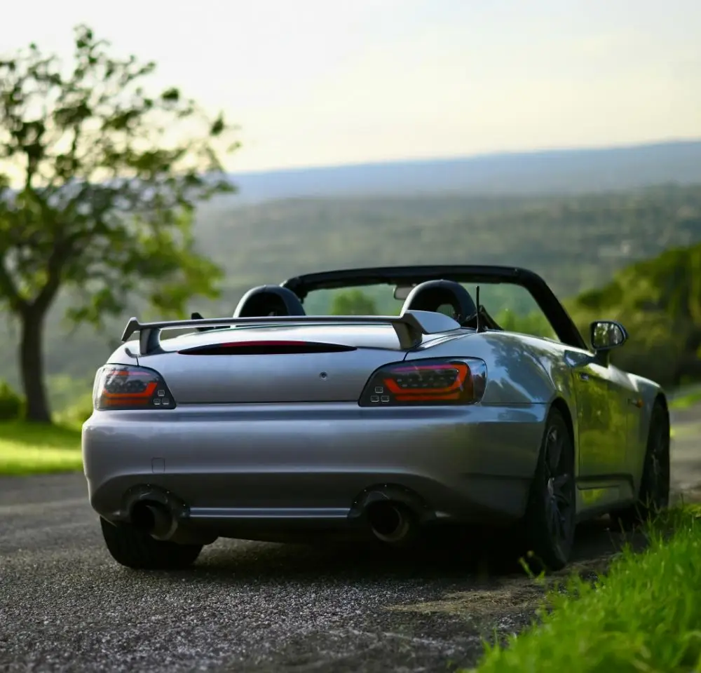 A silver Honda S2000 convertible sports car is parked on a scenic road with lush greenery and trees, overlooking a distant landscape under a clear sky—an ideal setting to consider Honda S2000 insurance for your prized motor.