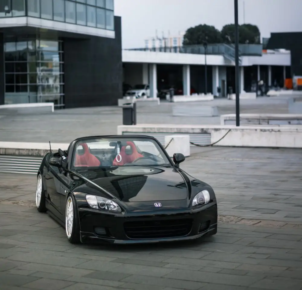 A black Honda S2000 convertible with red seats is parked on a modern, empty plaza in front of glass buildings on a cloudy day—perfect for those considering Honda S2000 cover or wanting to compare S2000 insurance rates.