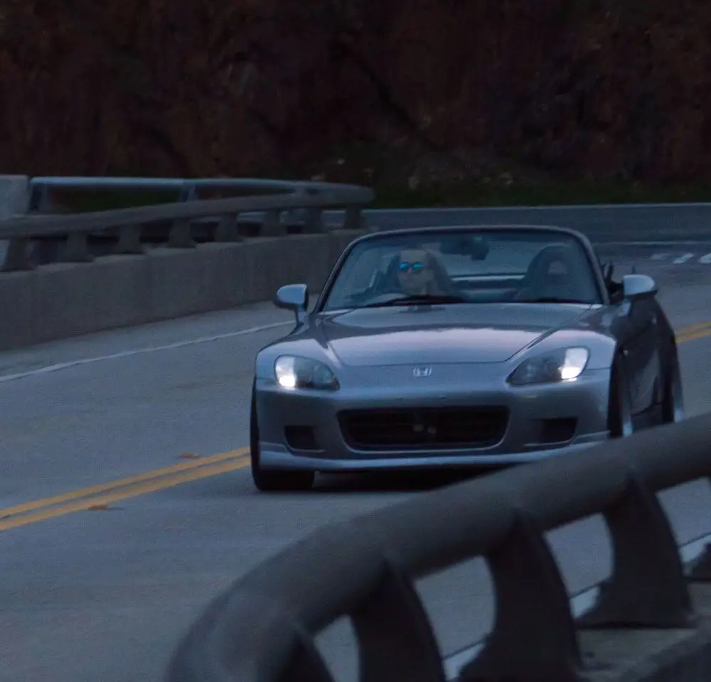 A silver Honda S2000 convertible sports car drives on a curved mountain road at dusk. The driver, wearing sunglasses, is visible behind the wheel. Concrete barriers line both sides of the road—a scene perfect for discussing S2000 insurance.