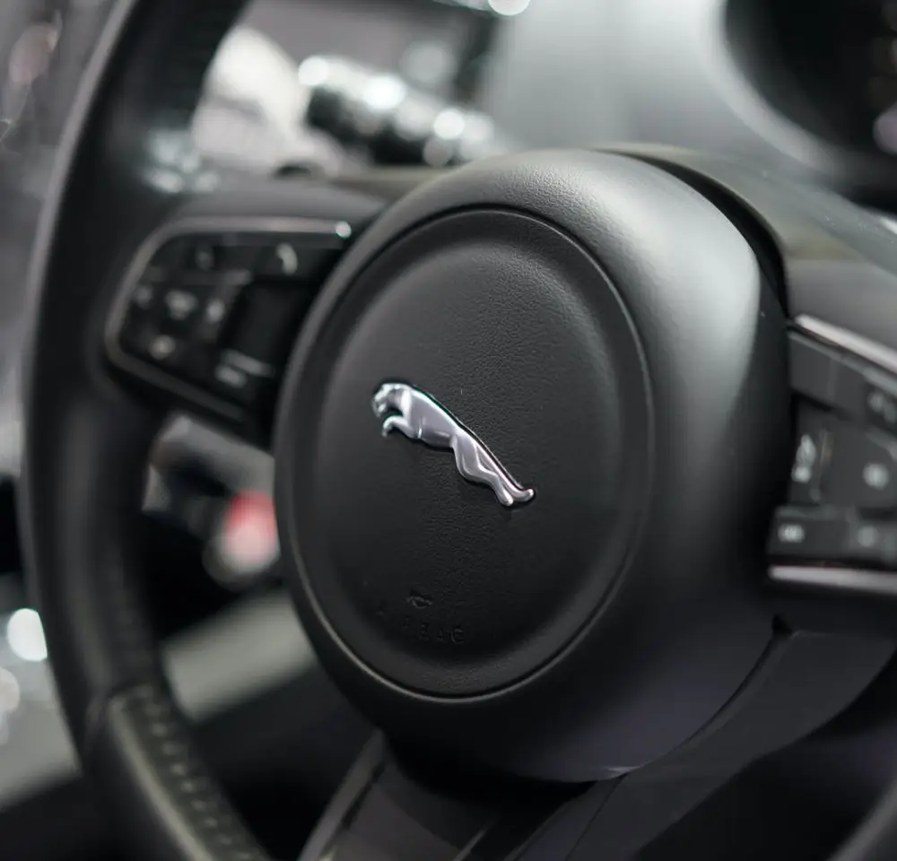 Close-up of a Jaguar car steering wheel, featuring the iconic silver leaping jaguar logo at the centre, with various control buttons and a discreet nod to Jaguar Insurance for comprehensive motor insurance protection.