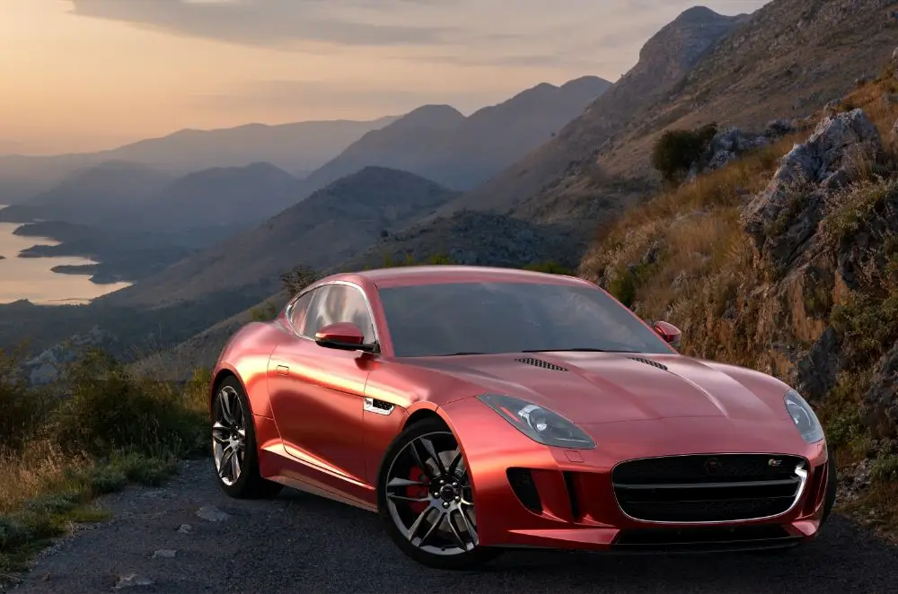 A shiny red sports car, covered by Jaguar Insurance, is parked on a mountain road at sunset, with rugged hills and a lake visible in the background.