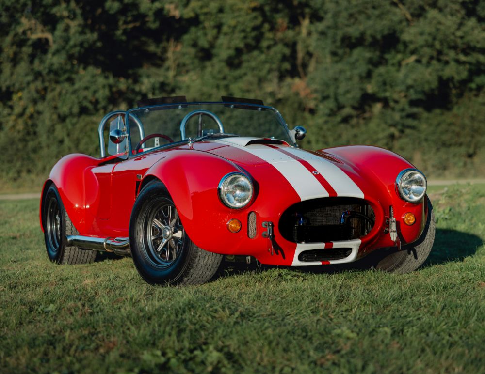 Classic red Shelby Cobra sports car with white racing stripes parked on a grassy field, featuring chrome side exhausts, black wheels, and a roll bar, set against a backdrop of dense green trees.