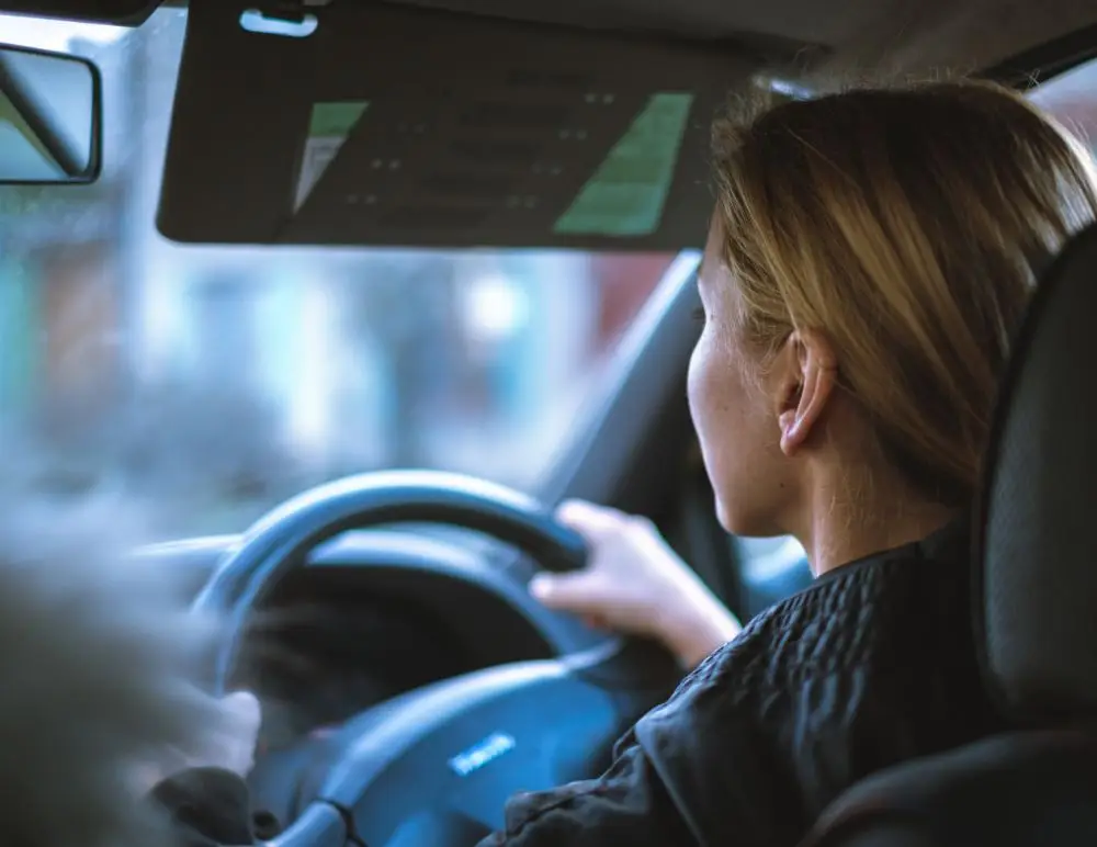 A woman with blonde hair is driving a car, seen from the passenger seat. She holds the steering wheel with both hands, focused on the road—a reminder of how important car insurance is for safety and peace of mind for women drivers.