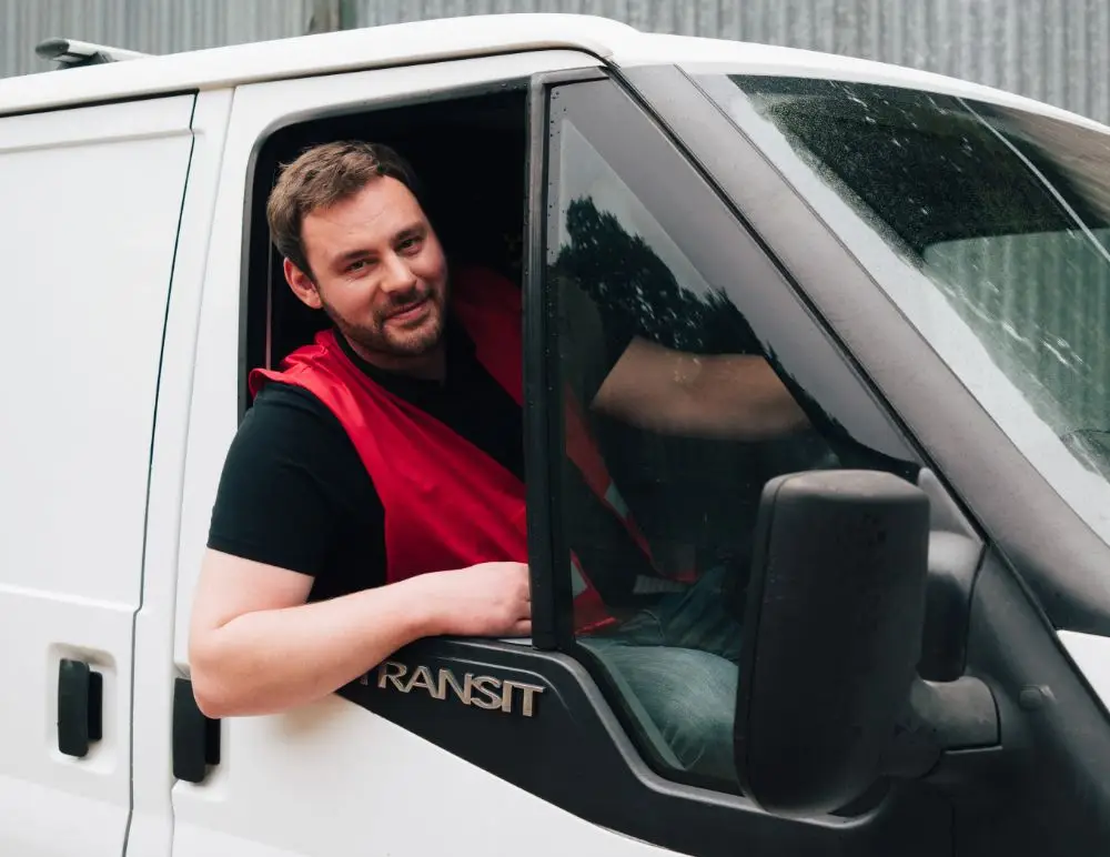 A man wearing a red safety vest sits in the driver’s seat of a white van, smiling at the camera with his arm resting on the open window.