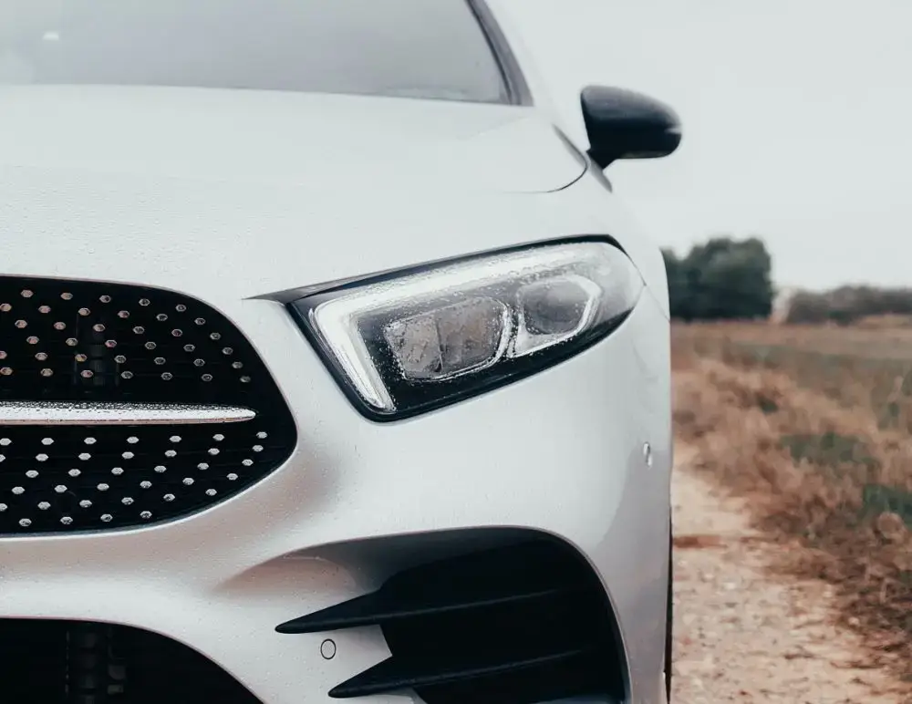 Close-up of the front left side of a white Mercedes-Benz, focusing on the headlamp, grille, and wing mirror, with an outdoor background featuring a dirt road and grassy area.