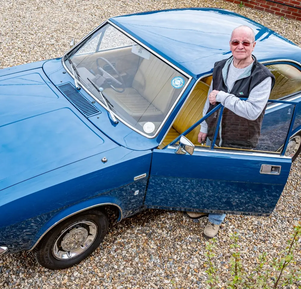 An older man wearing sunglasses stands beside an open door of a vintage blue Morris Marina parked on a gravel drive, smiling up at the camera—perhaps proud of both his classic car and his Morris Marina insurance.