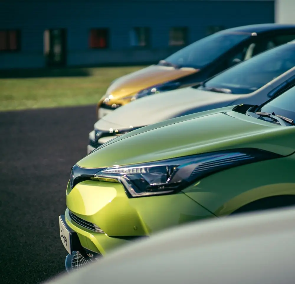 A row of modern cars in different colours, including green, gold, and silver, parked side by side on a paved surface with a building in the background—ideal options for those considering Toyota Insurance.