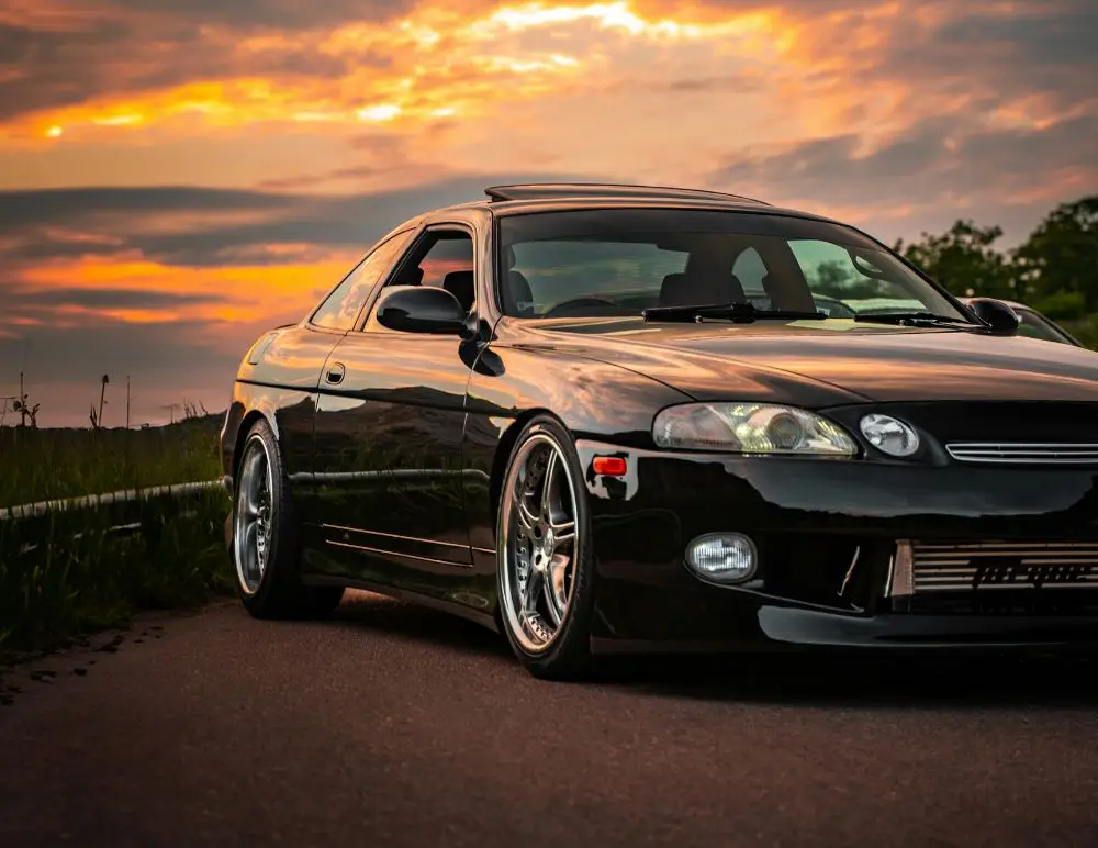 A sleek, black sports car with shiny chrome wheels is parked on a road at sunset, vibrant orange and yellow clouds overhead—a striking scene that highlights the appeal of securing reliable Toyota Soarer insurance. Only the front and side are visible.