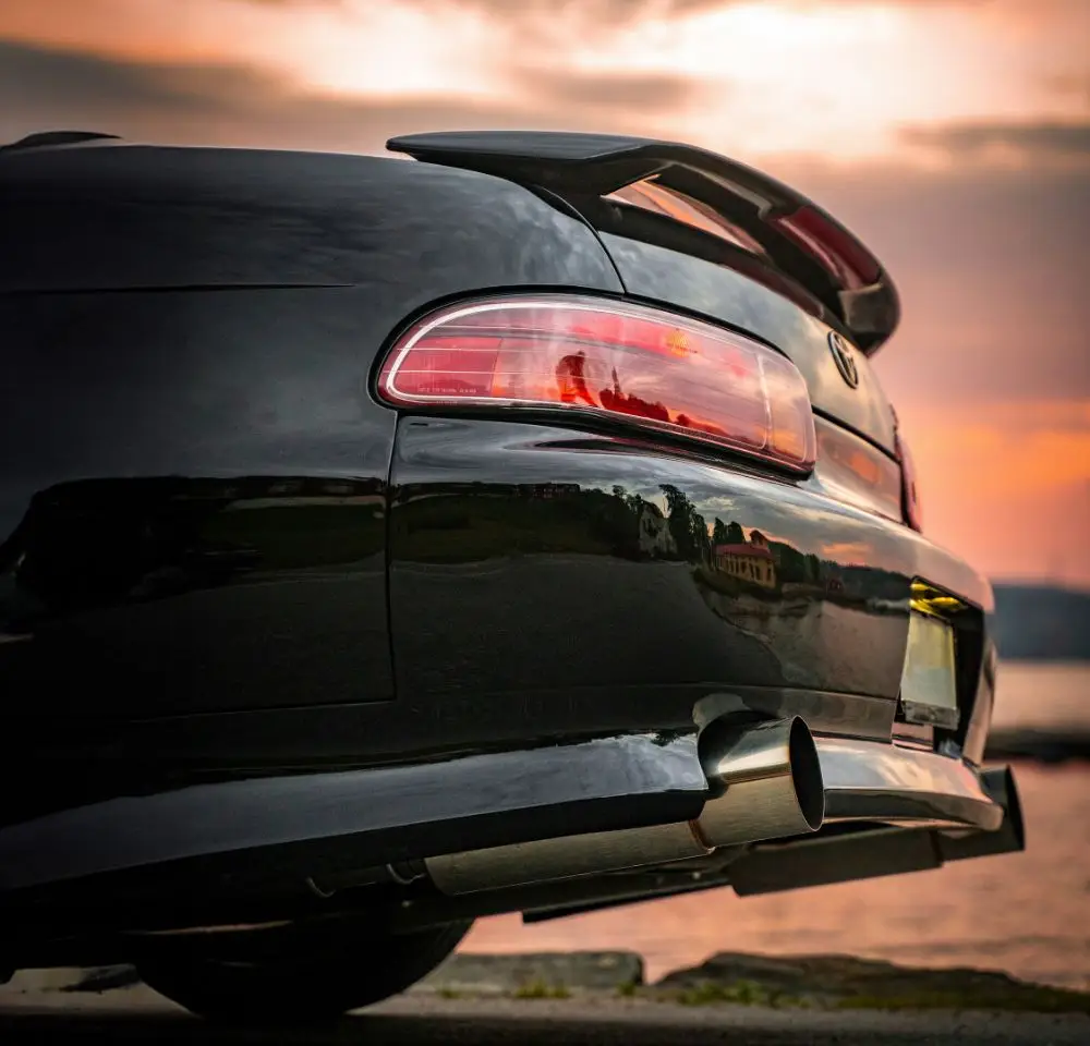 Close-up of the rear end of a black Toyota Soarer with a large spoiler, photographed at sunset near water, its shiny surface reflecting the scenic sky—perfect for those considering Toyota Soarer insurance.