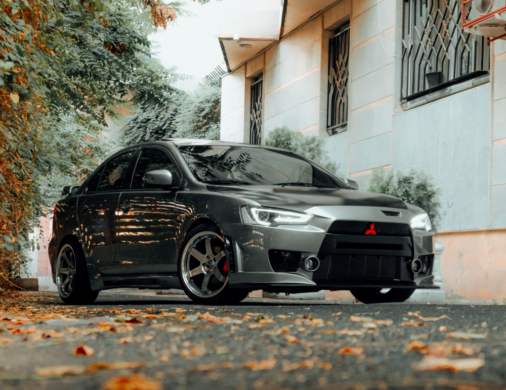 A grey Mitsubishi Lancer with custom alloys is parked on a tree-lined street, next to a beige building with barred windows. Fallen leaves are scattered on the ground—a perfect setting to showcase the need for reliable Mitsubishi Insurance.