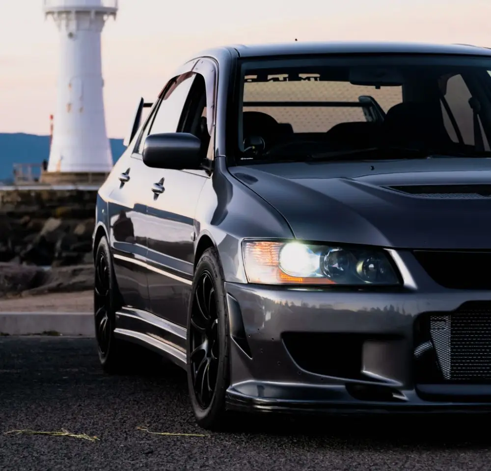 A close-up of a dark grey Mitsubishi Lancer with black wheels, parked near a white lighthouse on a rocky coast during sundown.