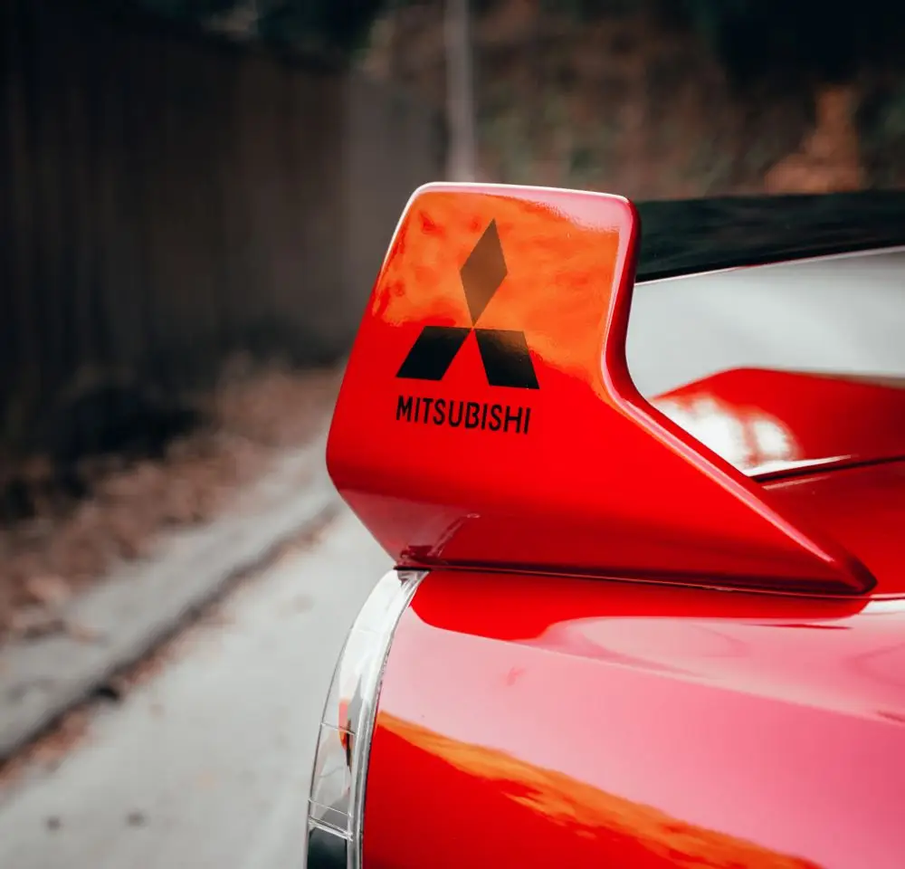 Close-up of a red Mitsubishi Lancer’s rear spoiler with the Mitsubishi logo and name, parked on a road with a blurred fence and foliage behind it—a reminder to keep your car insurance up to date.
