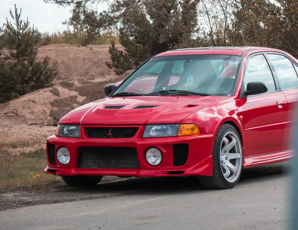 A bright red Mitsubishi Lancer Evolution parked on a road beside trees and sandy terrain, featuring sporty bodywork and silver alloy wheels—a true icon for any Mitsubishi Lancer fan considering car insurance.
