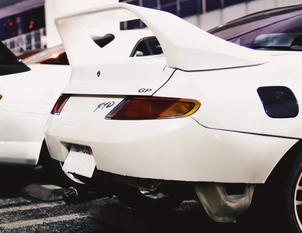 Close-up of the rear of a white Mitsubishi FTO with a large spoiler, GP and FTO badges, parked in a car park at night beside another white vehicle—a sleek sight for any Mitsubishi car insurance enthusiast.