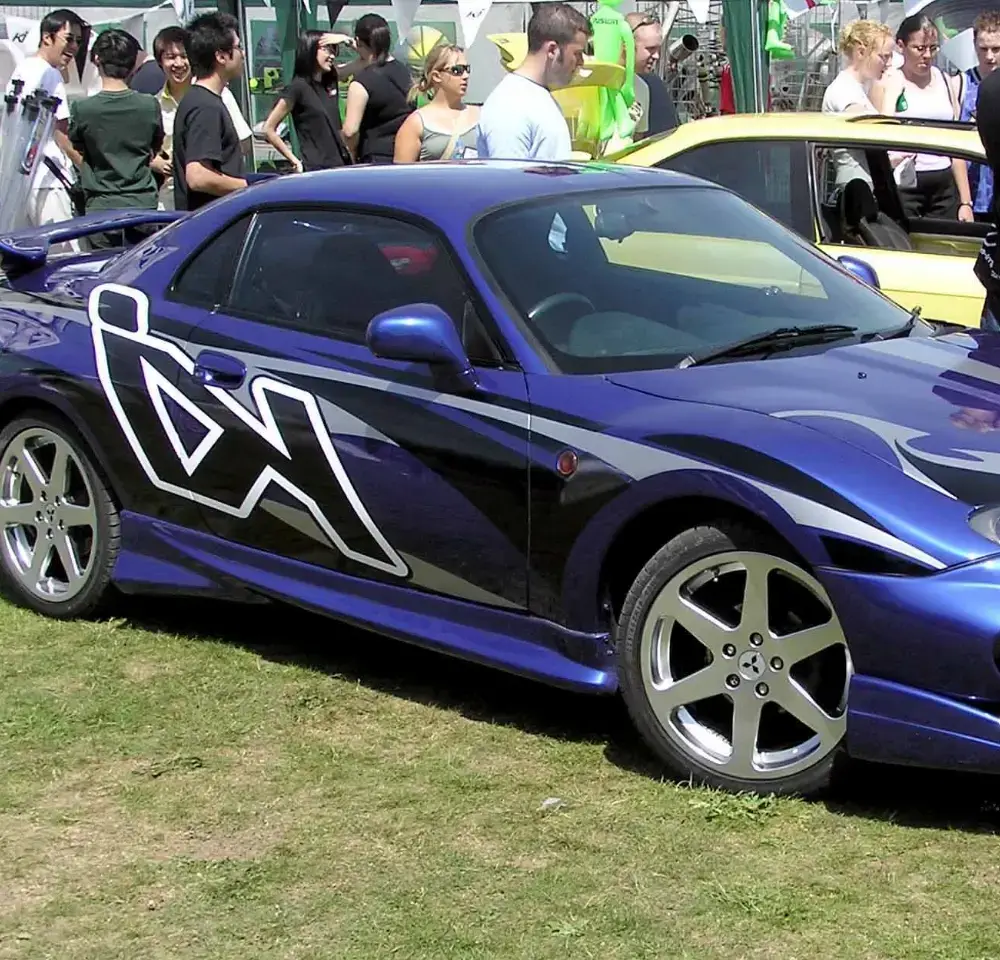 A blue Mitsubishi FTO sports car with a large white and black decal on the side is parked on grass at a car show, surrounded by people and other vehicles—it's an eye-catcher for any FTO insurance enthusiast.
