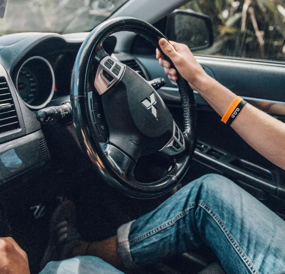 A person wearing jeans and a wristband is driving a Mitsubishi GTO, holding the steering wheel with their left hand. The car interior and dashboard are visible—a great reminder to consider Mitsubishi insurance for your motor.