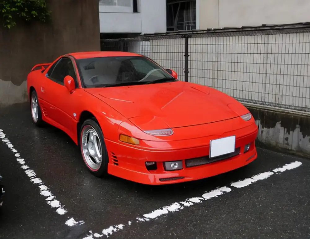 A bright red Mitsubishi GTO with pop-up headlamps is parked in a designated space on wet tarmac next to a metal fence and concrete wall, highlighting the allure of classic sports cars covered by Mitsubishi GTO insurance.