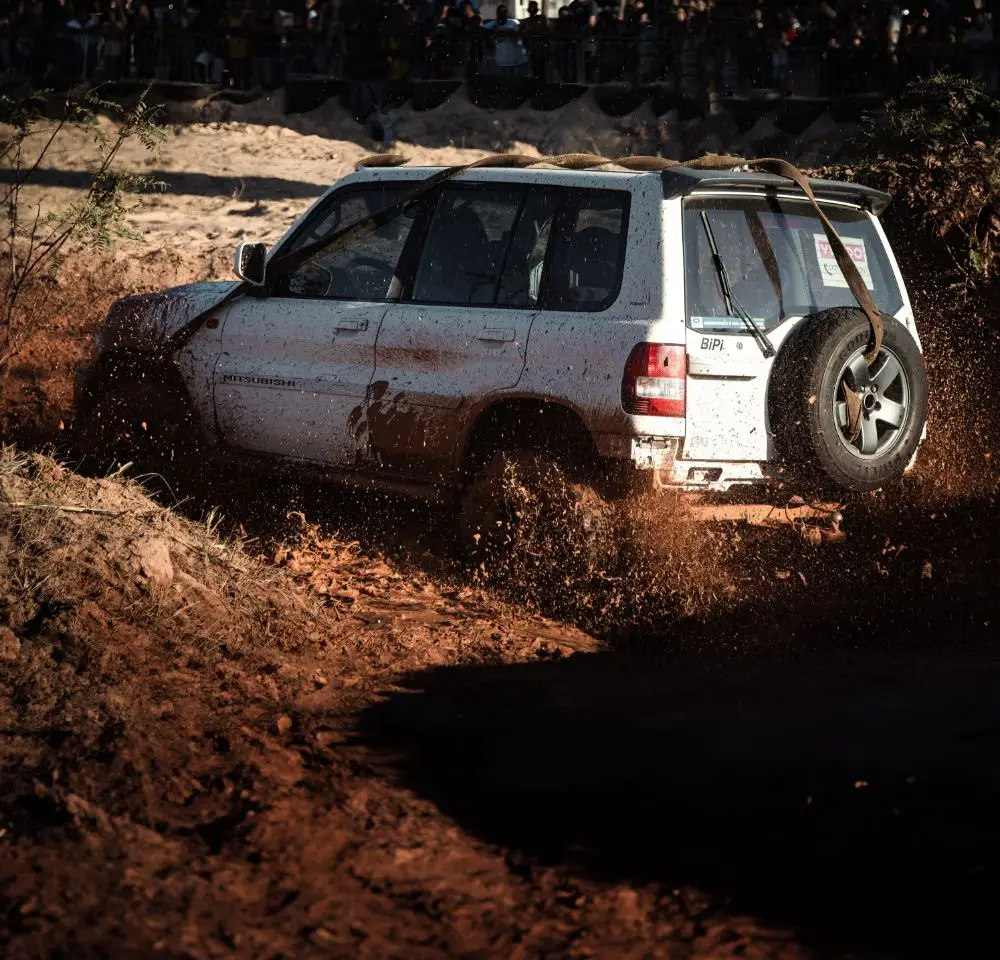 A white Mitsubishi SUV speeds through muddy terrain, splashing mud and water as it drives off-road. Mud flies up around the vehicle in a dynamic scene, with spectators visible in the blurred background—perfect for showing why Mitsubishi SUV insurance matters.