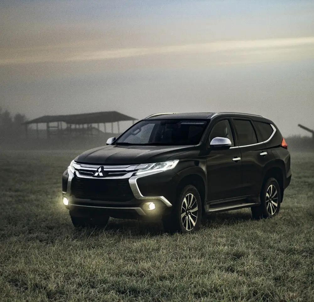 A black Mitsubishi SUV with headlights on is parked on a grassy field at dusk, mist swirling and barn silhouettes in the background—a scene perfect for those considering Mitsubishi Pajero insurance.