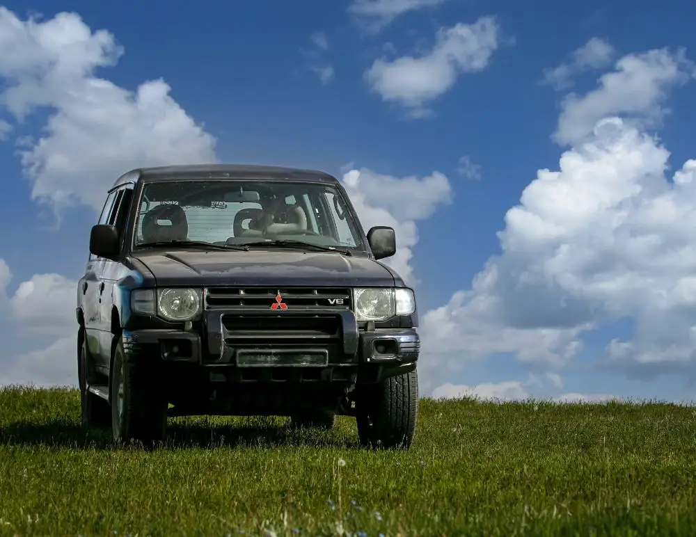 A black Mitsubishi SUV, ideal for Mitsubishi Pajero Insurance, is parked on a grassy field under a blue sky with scattered clouds. Sunlight highlights the green grass and the bright sky as the vehicle faces forwards.