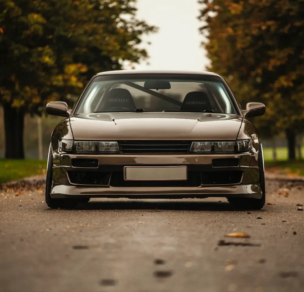 A lowered, dark-coloured Nissan Silvia with BRIDE racing seats is parked on a tree-lined street with fallen leaves and blurred trees in the background—showcasing a classic look perfect for Silvia insurance seekers.