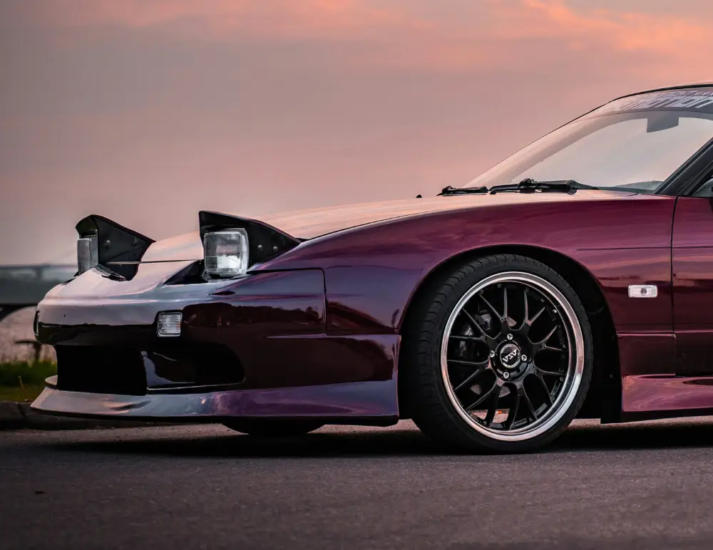 A maroon Nissan Silvia with pop-up headlamps and black alloy wheels is parked on the street during sunset. The front left side of the car is prominently visible, making a strong case for Nissan Silvia insurance.