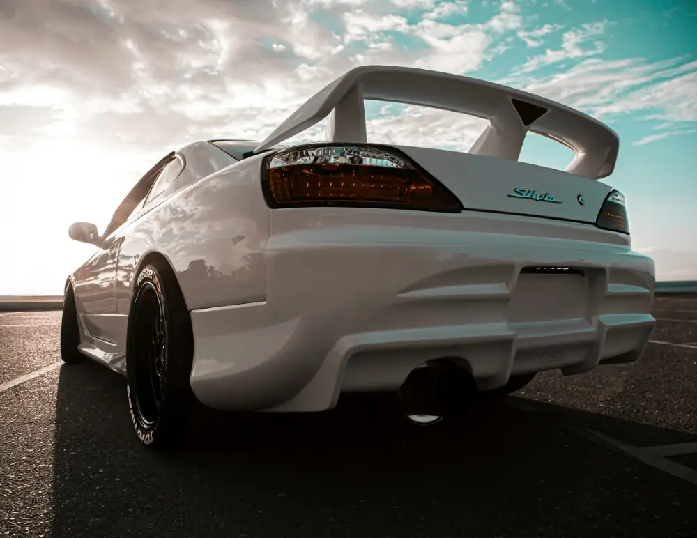 A low-angle view of a white Nissan Silvia sports car with a large rear spoiler parked on tarmac, the sun shining in the background and a partly cloudy sky.
