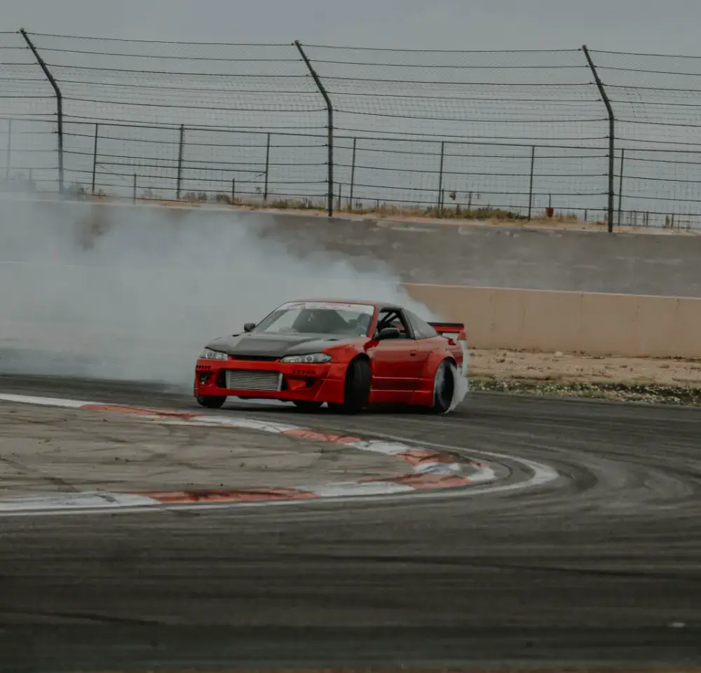 A red Nissan Silvia drifts around a track corner, creating a large cloud of tyre smoke behind it. The car is angled sideways, with barriers and a chain-link fence lining the track—perfect for showcasing why Nissan Silvia insurance matters.