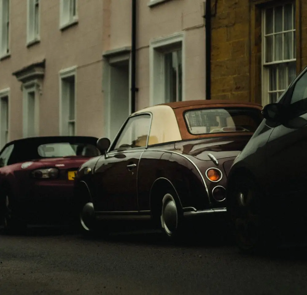 A vintage brown and cream Nissan Figaro is parked on a narrow street between two modern cars, with pastel-coloured buildings in the background. The dim lighting suggests early morning or evening—a perfect setting to capture classic style and consider Figaro insurance.
