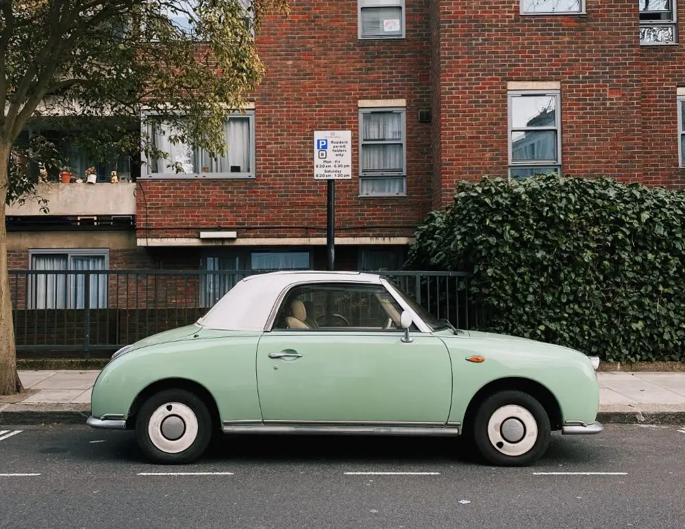 A vintage mint-green Nissan Figaro with a white roof is parked on a street in front of a brick block of flats, next to a tree and greenery, with a parking sign visible—perfect for showcasing classic style or Nissan Figaro insurance options.